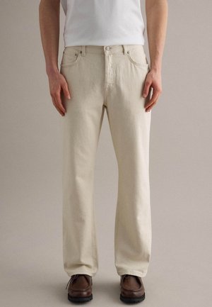 Persona che indossa jeans beige a gamba dritta, camicia bianca infilata e scarpe stringate marroni, in piedi davanti a uno sfondo neutro e semplice.