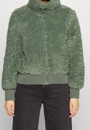 Persoon draagt een groene, zachte zip-up jas met geribde manchetten en zoom, gecombineerd met een donkere broek tegen een effen achtergrond.