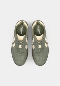 Grüne Wildleder-Sneaker mit beigen Akzenten und Schnürsenkeln, die ein Vintage-Design, eine strukturierte Oberfläche und eine braune Gummisohle aufweisen.