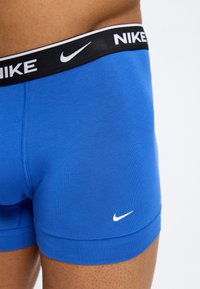 Κοντινό πλάνο σε μπλε boxer briefs της Nike με μαύρη ζώνη μέσης που φέρει λευκό λογότυπο Nike και swoosh στη ζώνη μέσης και στο πόδι.