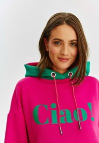 Jasnoróżowa, oversize'owa bluza z kapturem z zielonymi akcentami, na której dużą zieloną czcionką widnieje napis "Ciao!". Posiada regulowane sznurki oraz metalowe końcówki.