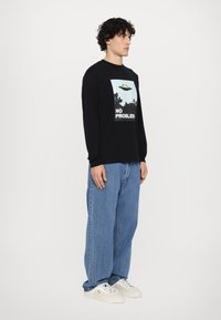 Black long-sleeve shirt featuring a graphic print with a UFO and text "NO PROBLEMS," paired with loose-fitting blue denim jeans and white sneakers.
