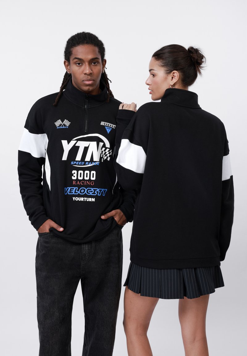 Black zip-up sweatshirt featuring white and blue prints and accents. The model wears dark jeans. The second model has a pleated black skirt.