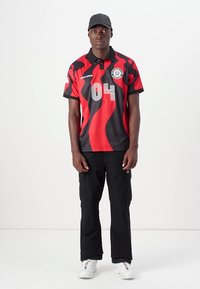 Red and black polo shirt with wavy pattern, featuring a collared neckline and "04" numeric detail. Paired with black cargo pants and white sneakers.