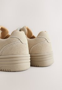Baskets en suède beige avec une finition texturée, talon arrondi et accents marron clair. Présentent des coutures contrastées et une semelle en caoutchouc.