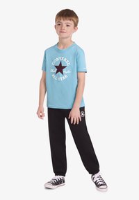 Svijetloplava majica s grafikom tamnocrvene zvijezde i bijelim tekstom, uparena s crnim trenirkom i crnim platnenim tenisicama.