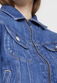 Denim jacket in blue, featuring a zip closure, pointed collar, two front pockets, and contrasting stitching details. Soft texture.