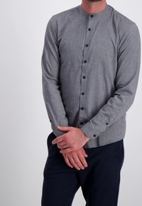 Harmaa nappikauluspaita, jossa on pyöreä pääntie, pitkät hihat ja mustat napit. Kangas näyttää olevan teksturoitu, hienovaraisella kuviolla. Yhdistetty tummiin pantsuihin.