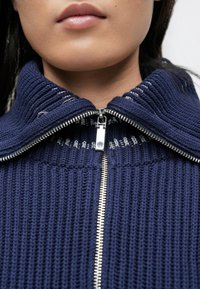 Tumesinine lukuga kardigan soonilise tekstuuriga ja kõrge kraega. Hõbedane tõmblukk ja hele õmbluse detail krae servas.