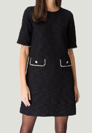 Femme portant une robe noire texturée à manches courtes avec des poches décoratives ornées de boutons argentés et de bordures blanches.