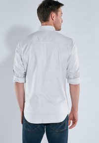 Weißes kariertes Hemd mit hochgekrempelten Ärmeln, ausgestattet mit einem Button-Down-Kragen und einem abgerundeten Saum. Der Stoff ist strukturiert und leicht.