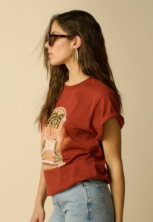 Frau mit langen braunen Haaren, die Schildpatt-Sonnenbrillen, goldene Kettenohrringe, rostfarbenes Grafik-T-Shirt und hellblaue Jeans trägt, blickt nach links.