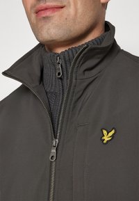 Uomo che indossa una giacca zip-up grigio scuro con colletto alto e logo di un uccello giallo sul petto sopra un maglione scuro a maglia con colletto a zip.