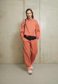 Lososově zbarvená zkrácená mikina a odpovídající tepláky s černou vrstvou pod nimi. Obsahuje logo Nike a má volný střih. Růžové tenisky.