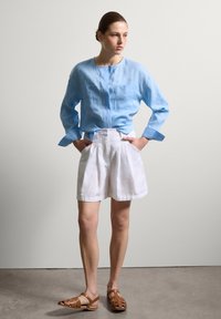 Hellblaues Leinenhemd mit langen Ärmeln und zwei Brusttaschen, kombiniert mit weißen Leinen-Shorts und braunen Riemchensandalen. Glatte Textur.