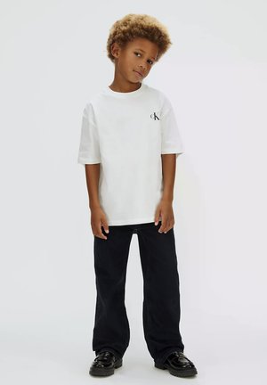 Giovane ragazzo con capelli ricci che indossa una t-shirt bianca Calvin Klein, pantaloni neri a gamba larga e scarpe nere, in piedi su sfondo bianco.