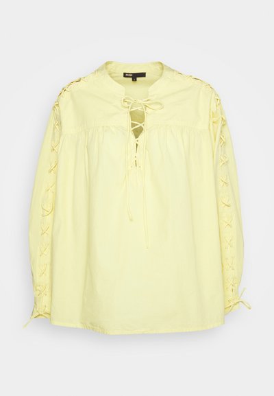 Blouse en coton jaune avec un col rond, un devant à lacets et un détail deçoratif en laçage le long des manches. Présente un tissu froncé pour plus de volume.