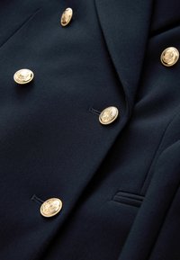 Navy blue, dwurzędowa kurtka z wytłaczanymi złotymi guzikami, gładka tkanina, ostre klapy i detal kieszeni z przodu.