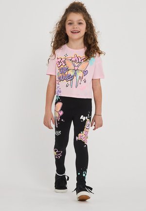 Maglietta cropped rosa con grafica colorata di farfalla, abbinata a leggings neri con stampe e disegni vivaci. Scarpe sportive nere.