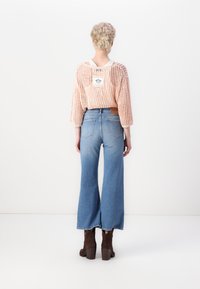 Könnyű, nyitott kötésű, barack színű pulóver a hátsó részen címkével, párosítva magas derekú, trapéznadrággal kék színben és barna sarkú csizmával.