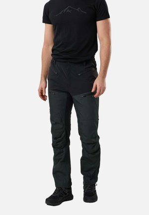 Uomo che indossa una maglietta nera con logo di montagna e pantaloni outdoor grigio scuro con tasche con zip e scarpe da trekking nere.