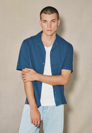 Joven con el cabello corto, vistiendo una camisa azul de manga corta sobre una camiseta blanca y pantalones vaqueros azul claro, de pie frente a un fondo beige.