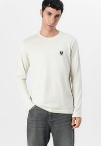 Long-sleeve cream shirt made of soft fabric, featuring a small black logo patch on the chest and a rounded crew neckline.