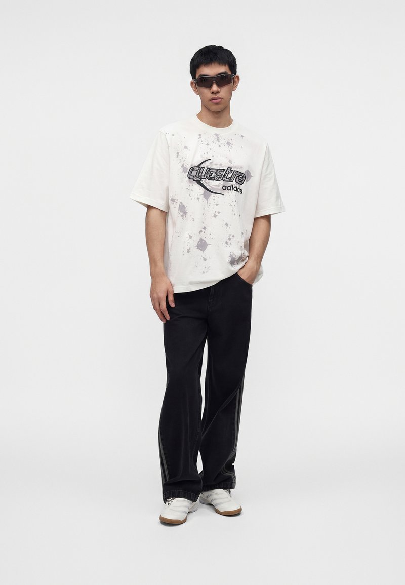 Giovane uomo che indossa una t-shirt bianca Adidas Questar, pantaloni neri a gamba larga, scarpe da ginnastica bianche e occhiali da sole scuri, in piedi con una mano in tasca.