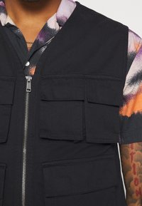 Czarna kamizelka bez rękawów z wieloma przednimi kieszeniami, zapinana na zamek błyskawiczny, wykonana z wytrzymałej tkaniny. Noszona na wzorzystej koszuli z krótkim rękawem.