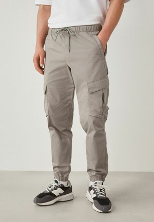 Man wearing light gray cargo pants with drawstring waist and elastic cuffs, paired with black and white sneakers standing on gray floor.