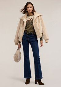 Palton din blană artificială bej cu un guler căptușit, pulover verde cu model, blugi negri evazați, și botine maro cu toc. Ține o geantă din pluș bej.