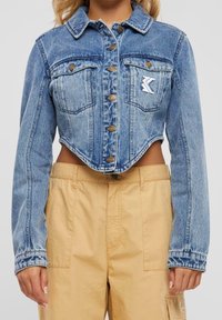 Mujer vistiendo una chaqueta de mezclilla azul recortada con botones dorados y pantalones cargo de tiro alto en color beige, mostrada desde el cuello hasta las caderas.