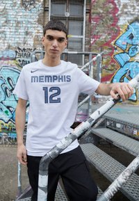T-shirt blanc à manches courtes portant "MEMPHIS" et "12" en bleu marine, associé à un pantalon cargo noir, devant un mur couvert de graffiti.