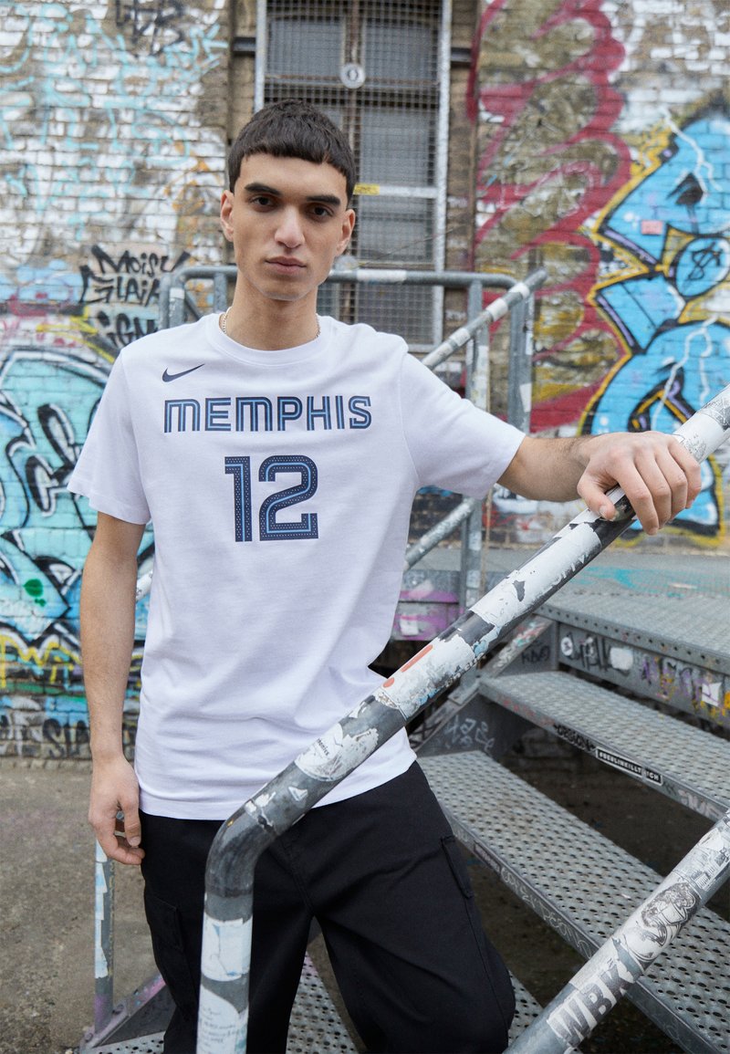 T-shirt blanc à manches courtes portant "MEMPHIS" et "12" en bleu marine, associé à un pantalon cargo noir, devant un mur couvert de graffiti.