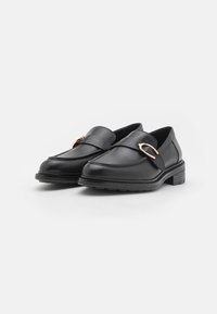 Mocasines de piel negra con un bajo tacón cuadrado, que presentan una hebilla decorativa en tono dorado y detalles de costura a lo largo del empeine. Suela de goma texturizada.