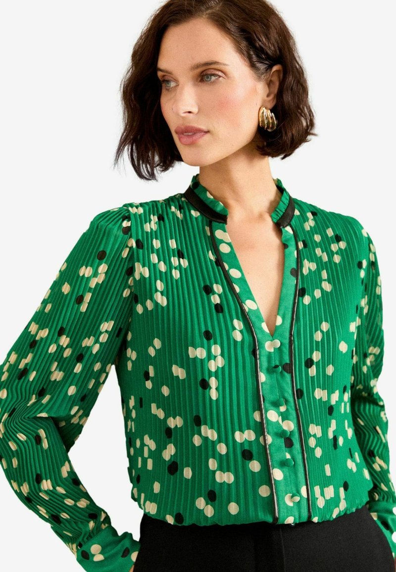 Blouse plissée verte avec des pois noirs et crème, présentant un design sans col et un devant à boutons, rehaussée d'une bordure noire.