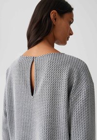 Une femme aux longs cheveux noirs porte un haut texturé gris et noir à motifs, avec une petite fente arrière et une fermeture à boutons près de l'encolure.