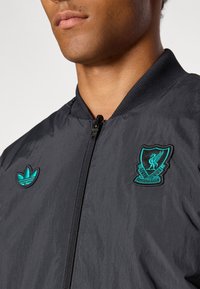 Μαύρο μπουφάν τύπου bomber με κεντημένα τιρκουάζ λογότυπα της Liverpool FC και της Adidas. Εμπρόσθιο φερμουάρ με μανσέτες και γιακά ριπ. Ελαφρύ υλικό.