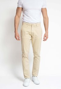 Hombre que lleva pantalones beige ajustados, camiseta blanca y zapatillas blancas, de pie frente a un fondo blanco plano.