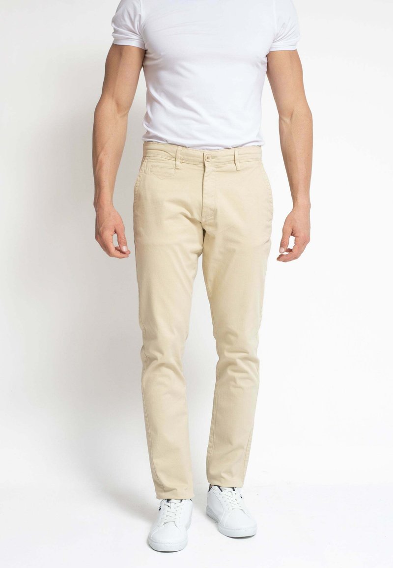 Hombre que lleva pantalones beige ajustados, camiseta blanca y zapatillas blancas, de pie frente a un fondo blanco plano.