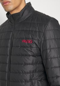 Svart pufferjacka med kviltad design, hög krage och en synlig röd "HUGO"-logotyp på bröstet. Har en framdragkedja och sidofickor.