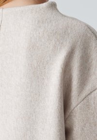 Vêtement en laine beige clair avec une surface douce et texturée et un col montant. Présente une coupe ample et d écontractée ainsi que des détails de coutures subtiles.