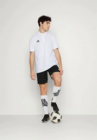 Λευκή polo μπλούζα με γιακά και μαύρο λογότυπο Adidas, μαύρες βερμούδες, ριγέ μαύρες και άσπρες κάλτσες και μαύρα παπούτσια ποδοσφαίρου, που στέκονται πάνω σε μια μπάλα ποδοσφαίρου.