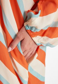 Robe multicolore avec des rayures diagonales orange, crème et bleu clair. Présente un poignet élastique et une poche latérale. Texture de tissu lisse.