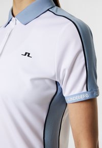 Vit polo med en blå ribbad krage, svarta sidostråk och en svart logotyp på bröstet. Accentuerad med "J.LINDEBERG" på ärmen.