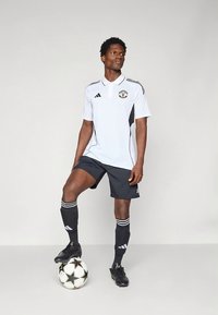 Vit fotbollströja med svarta detaljer, kombinerad med svarta shorts och strumpor. Svarta fotbollsskor och en fotboll med stjärndesign på marken.