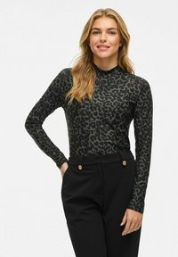 Jonge vrouw in een lange mouwtop met donkerluipaardprint en zwarte broek met gouden knopen, staand met één hand in haar zak, lachend.