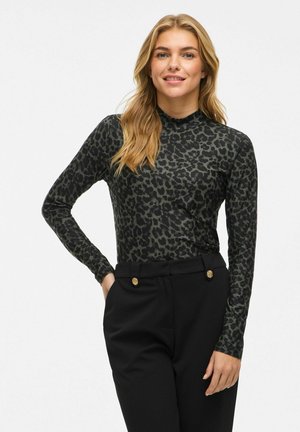 Jonge vrouw in een lange mouwtop met donkerluipaardprint en zwarte broek met gouden knopen, staand met één hand in haar zak, lachend.
