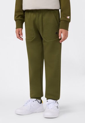 Pantalon de survêtement vert olive avec une taille élastique, des poches avant et un petit logo noir d'un côté. Associé à des baskets blanches.