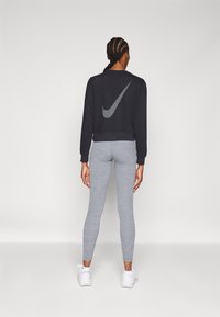 Črni skrajšani pulover s belim logotipom Nike na hrbtni strani, v kombinaciji s svetlo sivimi leginami in belimi supergami. Gladke teksture povsod.